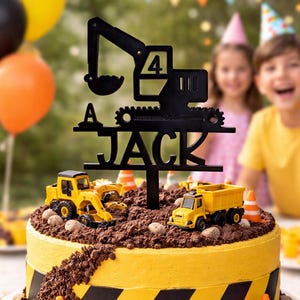 May include: A birthday cake decorated with a construction theme. The yellow cake has black hazard stripes and is topped with toy construction vehicles, chocolate 'dirt,' and a black topper that reads 'A JACK 4'. Orange traffic cones and balloons are in the background.