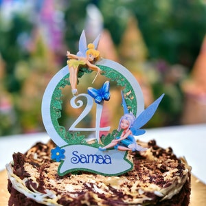 May include: A chocolate cake with a green and white fairy themed cake topper. The topper features two fairies, a butterfly, and the number 24. The cake is decorated with a blue flower and the name "Samaa" is written on the topper.