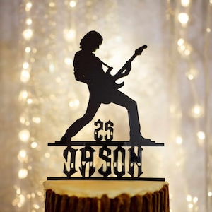 May include: Black silhouette of a person playing a guitar with the number 25 and the name Jason below. The silhouette is on a cake.