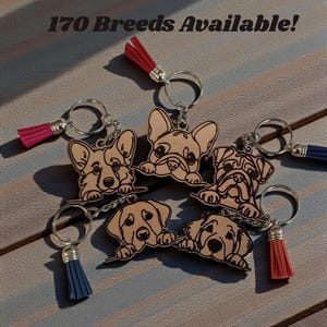 May include: Keychains featuring various dog breeds, each with a wooden cutout design. The keychains have silver rings and colorful tassels in red, pink, and blue. The text "170 Breeds Available!" is displayed at the top of the image.