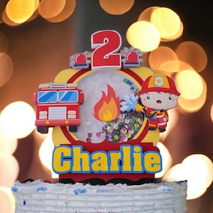 May include: A red, yellow, and blue cake topper with a fire truck, a fireman, a flame, and the number 2. The topper also has the name "Charlie" on it.