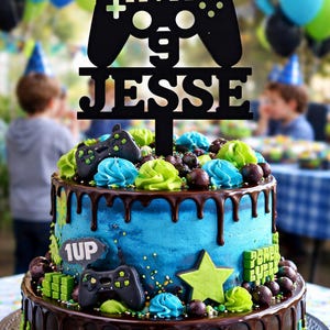 May include: A two-tiered birthday cake with blue frosting, chocolate ganache, and decorative elements. The cake is adorned with a black controller-shaped topper that reads "LEVEL 9 JESSE." Additional decorations include green and blue frosting swirls, chocolate candies, and a green star.