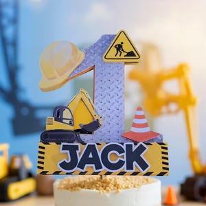 May include: A yellow and black construction themed cake topper with a number one, a toy excavator, a hard hat, a construction sign, and a traffic cone. The topper also has the name "JACK" on it.