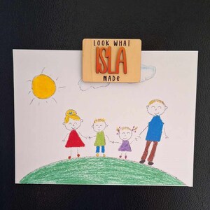 May include: A wooden sign with the text "Look What Isla Made" in orange letters on a light brown background. The sign is attached to a white piece of paper with a drawing of a family of four on it. The drawing is done in crayon and shows a sun, clouds, and a green hill.