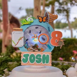 May include: A blue and green cake topper with a magnifying glass, a jar with a firefly, a butterfly, a net, and the number 8. The topper also has the name "Josh" on a wooden sign.