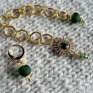 May include: Gold-toned jewelry set featuring a bracelet and an earring. The bracelet is made of linked circles. The earring and bracelet have green beads and a sunburst design with a green center stone. The jewelry is set against a white knitted background.