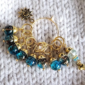 Könnte beinhalten: Eine Sammlung goldfarbener Maschenmarkierer mit blauen und goldenen Perlen. Die Maschenmarkierer sind an einem goldenen Ring befestigt und haben einen Sonnenanhänger. Die Perlen haben ein strukturiertes, craquelé Aussehen.