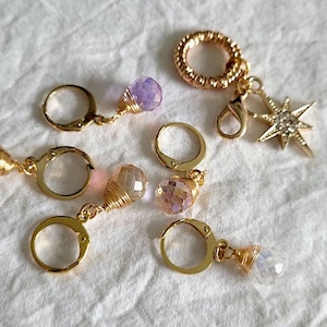 Pode incluir: Um conjunto de seis brincos dourados com pingentes de cristal. Os pingentes são em tons de roxo, rosa e transparente. Os brincos são presos a uma corrente dourada com um pingente de estrela.