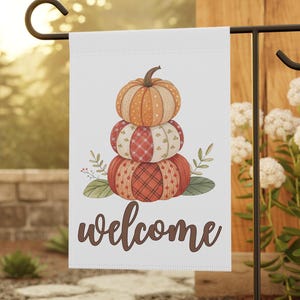 May include: A white garden flag featuring a stack of three pumpkins in shades of orange and red, with a brown stem. The word "welcome" is written in a brown script font below the pumpkins. The flag is displayed on a black metal garden stake.