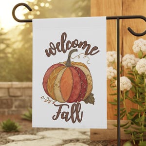 May include: White garden flag with a watercolor pumpkin design. The pumpkin is in shades of orange, red, and yellow with the words "welcome Fall" in brown script. The flag is displayed on a black metal stand.