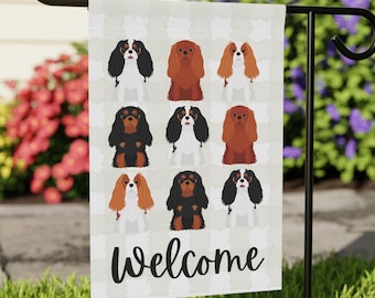 May include: A welcome garden flag with a white background and a pattern of Cavalier King Charles Spaniels in various colors. The flag says "Welcome" in black cursive lettering.