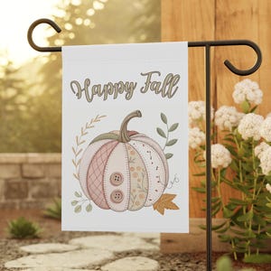 May include: A white garden flag with the text "Happy Fall" in a script font. The flag features a pumpkin illustration with pink, tan, and green accents, and two brown buttons. The flag is displayed on a black metal stand.