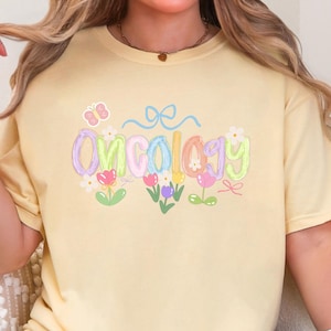 May include: A light yellow t-shirt with the word "Oncology" in pastel colors, decorated with flowers and a butterfly. The shirt is a short-sleeved, crew neck style.