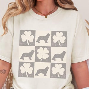 Puede incluir: Una camiseta blanca con un gráfico gris de cuatro perros y cuatro tréboles. Los perros están todos en posición sentada y los tréboles tienen un diseño de trébol de cuatro hojas.