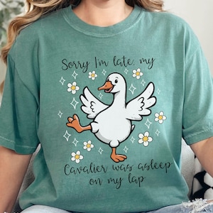 Puede incluir: Una camiseta verde mar con un dibujo de un pato de dibujos animados y el texto "Sorry I'm late, my Cavalier was asleep on my lap." El pato es blanco con patas naranjas y un pico amarillo, rodeado de flores blancas.