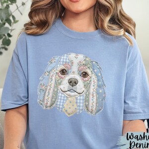 May include: Light blue t-shirt featuring a patchwork design of a Cavalier King Charles Spaniel dog. The dog's face is detailed with floral and gingham patterns in shades of pink, blue, and white. The text "Washed Denim" is visible on the lower right.