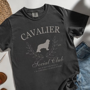 Puede incluir: Una camiseta gris oscuro con un diseño de estilo vintage que presenta una silueta de un perro Cavalier King Charles Spaniel, el texto "Cavalier Social Club" y el texto "Est. 2006".