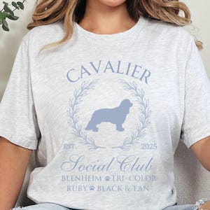 Puede incluir: Una camiseta gris con una silueta azul de un perro Cavalier King Charles Spaniel dentro de una corona con el texto "Cavalier Social Club" y "Est. 2025" debajo de la corona. El texto "Blenheim, Tri-Color, Ruby, Black & Tan" está debajo del nombre del club.