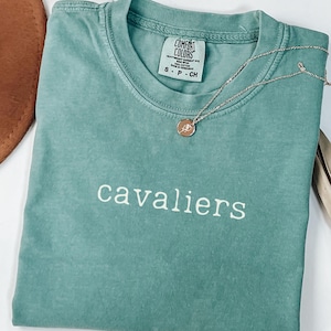 Puede incluir: Una camiseta azul verdosa con la palabra "cavaliers" bordada en blanco en la parte delantera. La camiseta está hecha de una tela suave y cómoda y tiene un corte holgado.