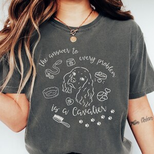 Puede incluir: Camiseta gris oscuro con el texto "la respuesta a cada problema es un Cavalier" y una ilustración blanca de un Cavalier King Charles Spaniel y artículos para perros. Estilo casual.