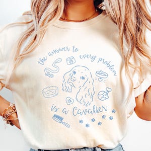Puede incluir: Camiseta color crema con una ilustración azul claro de un Cavalier King Charles Spaniel e iconos relacionados con perros. El texto "the answer to every problem is a Cavalier" está escrito en una fuente de escritura.