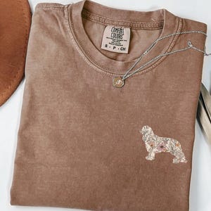 Puede incluir: Una camiseta marrón con una silueta de perro floral bordada en la parte delantera. La camiseta tiene cuello redondo y está hecha de una tela suave y cómoda. La etiqueta de la camiseta dice "Comfort Colors" y "S - P - CH".