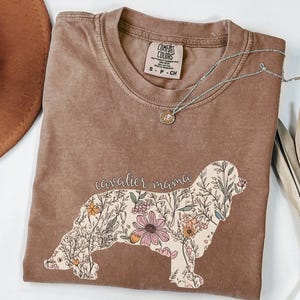 Puede incluir: Una camiseta marrón con un diseño floral de un Cavalier King Charles Spaniel. El texto "Cavalier mama" está impreso en la camiseta.