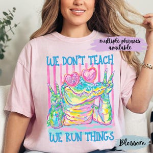 K&ouml;nnte beinhalten: Ein hellrosa T-Shirt mit einer bunten Grafik eines Alligators, der herzf&ouml;rmige Sonnenbrillen tr&auml;gt und mit beiden H&auml;nden das Friedenszeichen zeigt. Der Text lautet WE DON'T TEACH WE RUN THINGS vor einem Hintergrund mit vertikalen rosa Streifen.