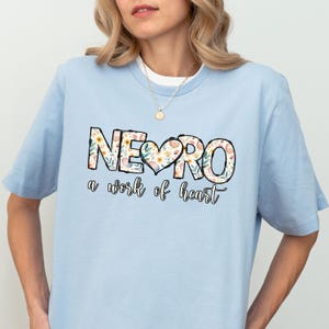 May include: Light blue t-shirt with a floral design spelling out "NEURO" with a heart in the middle and the text "a work of heart" below.