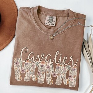 May include: A brown t-shirt with the text "Cavalier Mama" in white letters with a floral pattern. The shirt has a distressed look and is made of a soft, comfortable fabric.