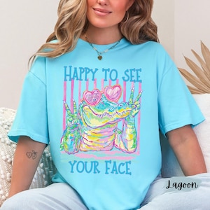 K&ouml;nnte beinhalten: Ein hellblaues T-Shirt mit einer bunten Grafik eines Alligators, der eine rosa herzf&ouml;rmige Sonnenbrille tr&auml;gt und mit beiden H&auml;nden das Friedenszeichen zeigt. Der Text HAPPY TO SEE und YOUR FACE ist in blauen Buchstaben gedruckt.
