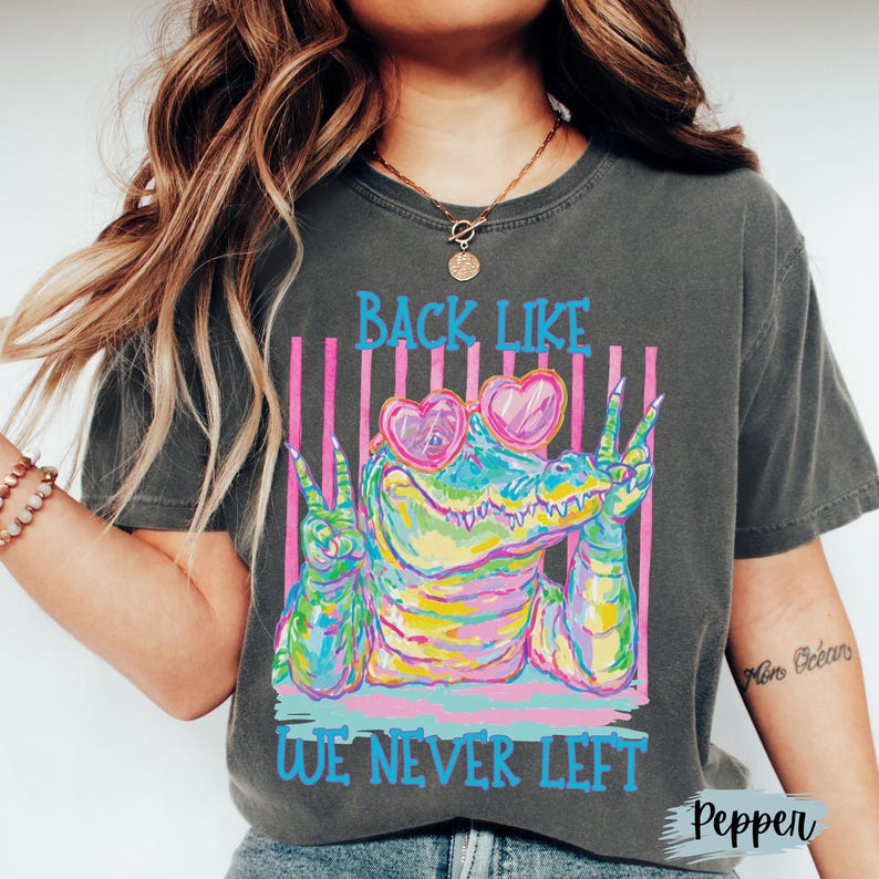 K&ouml;nnte beinhalten: Ein anthrazitfarbenes Kurzarm-T-Shirt mit einer bunten Grafik eines Alligators, der eine herzf&ouml;rmige Sonnenbrille tr&auml;gt und mit beiden H&auml;nden das Friedenszeichen zeigt. Der Text BACK LIKE WE NEVER LEFT steht in leuchtendem Blau dar&uuml;ber und darunter.