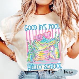 K&ouml;nnte beinhalten: Ein elfenbeinfarbenes T-Shirt mit einer bunten Grafik eines Alligators, der herzf&ouml;rmige Sonnenbrillen tr&auml;gt und mit beiden H&auml;nden Friedenszeichen macht. Der Text "GOOD BYE POOL" steht &uuml;ber dem Alligator und "HELLO SCHOOL" darunter.