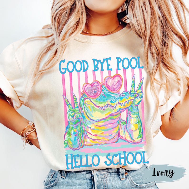 K&ouml;nnte beinhalten: Ein elfenbeinfarbenes T-Shirt mit einer bunten Grafik eines Alligators, der herzf&ouml;rmige Sonnenbrillen tr&auml;gt und mit beiden H&auml;nden Friedenszeichen macht. Der Text "GOOD BYE POOL" steht &uuml;ber dem Alligator und "HELLO SCHOOL" darunter.
