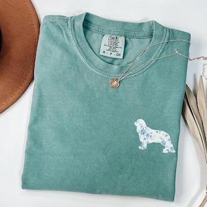 Puede incluir: Una camiseta verde agua Comfort Colors con un diseño floral de perro. Un collar con colgante dorado y un sombrero marrón también están en la imagen. La camiseta tiene una etiqueta pequeña con la marca y la talla.