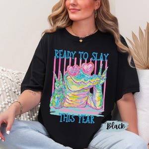 K&ouml;nnte beinhalten: Ein schwarzes T-Shirt mit einer bunten Grafik eines Alligators, der eine rosa herzf&ouml;rmige Sonnenbrille tr&auml;gt und mit beiden H&auml;nden das Friedenszeichen zeigt. Der Text READY TO SLAY und THIS YEAR ist in leuchtendem Blau gedruckt.