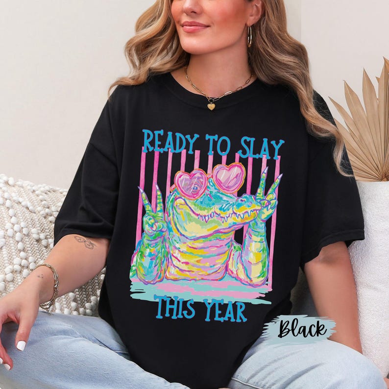 K&ouml;nnte beinhalten: Ein schwarzes T-Shirt mit einer bunten Grafik eines Alligators, der eine rosa herzf&ouml;rmige Sonnenbrille tr&auml;gt und mit beiden H&auml;nden das Friedenszeichen zeigt. Der Text READY TO SLAY und THIS YEAR ist in leuchtendem Blau gedruckt.
