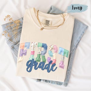 May include: Ivory-colored t-shirt with the words "FIRST grade" in a patchwork design. The letters are made of various patterned fabrics, including floral and gingham prints. A gold necklace rests on the shirt, which is folded on top of a pair of blue jeans.