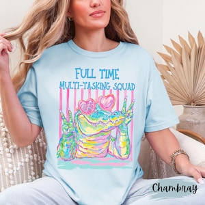 K&ouml;nnte beinhalten: Ein hellblaues T-Shirt mit Rundhalsausschnitt und einer Grafik eines bunten Alligators, der herzf&ouml;rmige Sonnenbrillen tr&auml;gt und mit beiden H&auml;nden das Friedenszeichen zeigt. Dar&uuml;ber steht der Text FULL TIME MULTI-TASKING SQUAD in Blau.