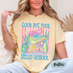 K&ouml;nnte beinhalten: Ein hellgelbes T-Shirt mit einer bunten Grafik eines Alligators, der herzf&ouml;rmige Sonnenbrillen tr&auml;gt und mit beiden H&auml;nden das Friedenszeichen zeigt. Der Text GOOD BYE POOL und HELLO SCHOOL steht in blauen Buchstaben vor einem rosa gestreiften Hintergrund.