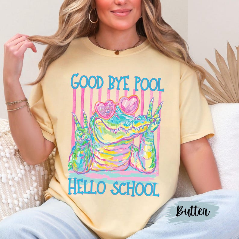 K&ouml;nnte beinhalten: Ein hellgelbes T-Shirt mit einer bunten Grafik eines Alligators, der herzf&ouml;rmige Sonnenbrillen tr&auml;gt und mit beiden H&auml;nden das Friedenszeichen zeigt. Der Text GOOD BYE POOL und HELLO SCHOOL steht in blauen Buchstaben vor einem rosa gestreiften Hintergrund.