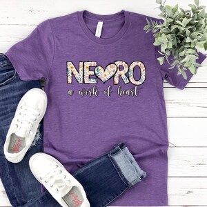 May include: Purple t-shirt with a floral design that spells out "NEURO" with a heart in the middle. The text below the design reads "a work of heart".