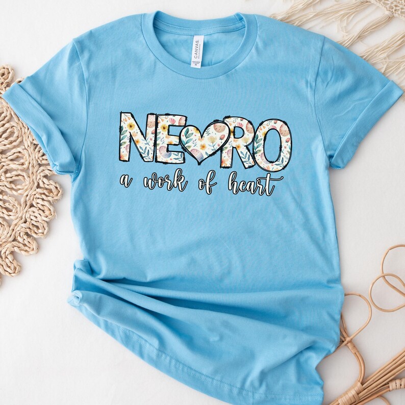 May include: A light blue t-shirt with a floral design that spells out "NEURO" with a heart in the middle. The text below the design reads "a work of heart".
