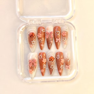 Puede incluir: Un juego de diez uñas artificiales en una caja de plástico transparente. Las uñas en forma de almendra presentan varios diseños, incluyendo purpurina, lazos, flores y personajes de dibujos animados en tonos naranja, rosa y blanco.