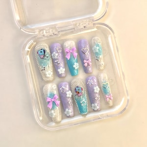 Puede incluir: Un juego de diez uñas postizas en forma de ataúd en una caja de plástico transparente. Las uñas presentan una variedad de diseños, que incluyen purpurina, lazos, corazones y personajes de dibujos animados en tonos de azul, morado y blanco. Algunas uñas tienen acentos florales.