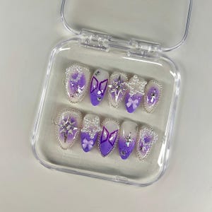 May include: A set of ten artificial nails in a clear plastic case. The nails feature a gradient of purple and white, adorned with silver glitter, star-shaped embellishments, and small bows. The nails are oval-shaped.