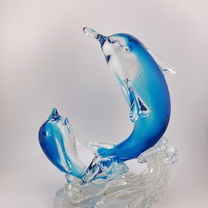 May include: A glass sculpture of two blue dolphins leaping out of the water. The dolphins are made of clear glass with blue accents. The sculpture is set on a clear glass base that resembles waves.