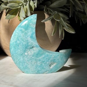 May include: A turquoise crescent moon-shaped crystal sculpture. The stone has a marbled appearance with white and light blue hues. The sculpture is displayed on a white surface, with a wooden vase and greenery in the background.