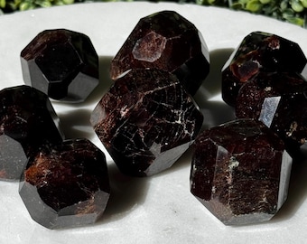 May include: A collection of seven dark red, faceted garnet crystals with natural inclusions and geometric shapes. Each stone displays a deep burgundy hue with polished surfaces that reflect light, showcasing the raw, angular structure of the minerals.