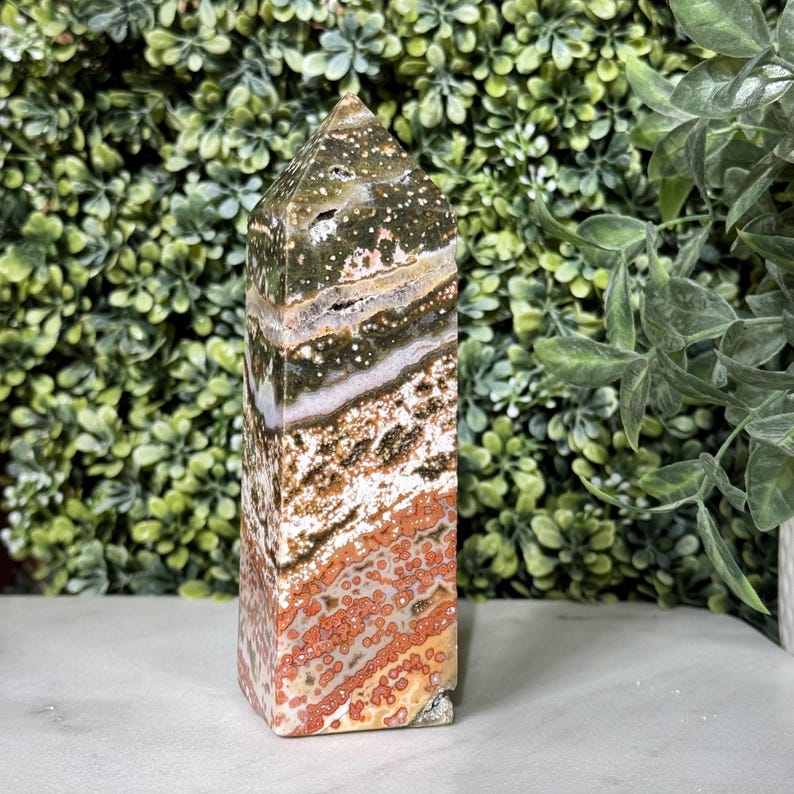 May include: A tall, rectangular Ocean Jasper crystal tower with a pointed top. The stone features a mix of colors, including green, white, brown, and red, with a speckled and banded pattern. The crystal is standing upright.